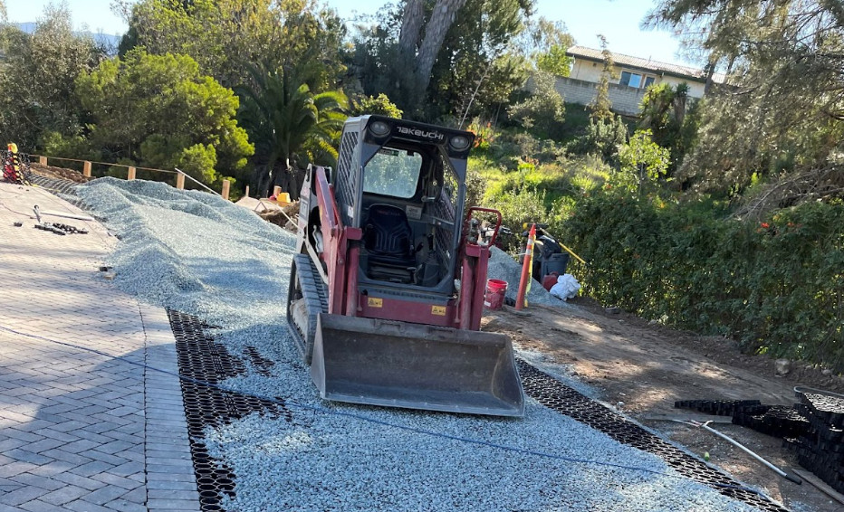 Compact loader spreading gravel over a driveway under construction.