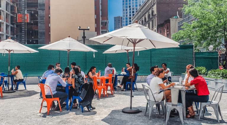 People dining outdoors at café tables with umbrellas in a city setting.