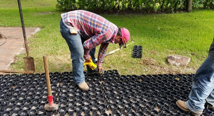 Contractor securing 24x24 plastic pavers in place over a prepared ground base for a permeable surface installation.