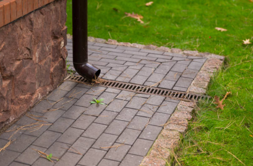 Downspout directing water into a channel drain along a paver walkway for home stormwater management.